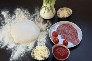 Pizzateig auf Mehl, Fenchelknolle mit Dill, geriebener Mozzarella, Fenchelsalami, Kirschtomaten und Tomatensoße für Pizza mit Fenchel und Fenchelsalami
