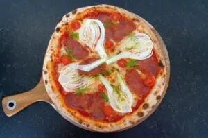 Pizza auf Holzbrett mit Salami, Tomatenscheiben, Fenchel und Kräutern auf dunklem Untergrund