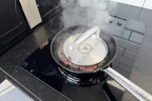 Smashburger-Pizza in Grillpfanne mit Deckel, Fleischpatty unter Edelstahldeckel auf heißer Pfanne auf Gasgrill gepresst