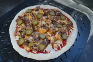 Smashburger-Pizza auf Grillrost: Pizzaboden mit Tomatensoße, Käse, gehacktem Rinderfleisch, Cheddar und Gürkchen
