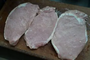 Drei rohe Schweineschnitzel mit Salz und Pfeffer gewürzt auf Holzbrett, Vorbereitung für Tonkatsu