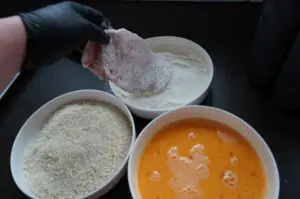 Hand mit Handschuh paniert rohes Schweineschnitzel mit Semmelbröseln, links Mehlschüssel, oben Ei, rechts fertige Panade