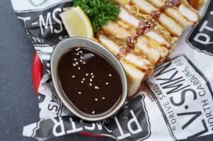 Schüssel mit dunkler Tonkatsu-Sauce mit Sesamkörnern neben gegrilltem Tonkatsu mit knuspriger Panade, Zitronenscheibe und frischer Petersilie