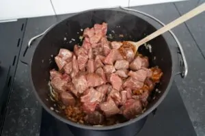 Rinderfleischwürfel werden in schwarzem Topf auf Grillfeld mit Holzlöffel gerührt, Zwiebeln angeröstet, Zubereitung Wiener Saftgulasch