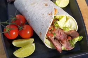 Flank-Steak-Wrap mit grünem Salat, Salsa und gegrilltem Flank Steak in Tortilla, dazu Limettenspalten und Strauchtomaten