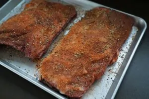 Zwei Wildschwein-Ribs auf weißem Tablett mit großzügiger Gewürzmischung aus Paprika und Kräutern eingerieben, Vorbereitung zum Grillen