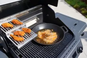 Knusprig frittierte Chicken Fingers in einer schwarzen Pfanne mit Öl auf einem Gasgrill, daneben vier weitere Finger auf dem Grillrost