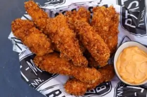 Knusprig panierte Hähnchen-Finger mit goldbrauner Kruste auf schwarzweißem Papier mit Sauce in Schälchen daneben