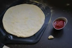 Pizza Marinara - Original-Rezept aus Neapel 7 Pizzateig auf schwarzem Grillblech, daneben Schüssel mit Tomatensoße und Knoblauch