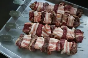 Rindfleischspieße mit Camembert und Bacon 10 Vier Rindfleischspieße mit Bacon umwickeltem Rindfleisch und Camembert-Würfeln auf weißem Tablett mit Grillspießen, rohe Zutaten vor dem Grillen