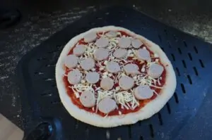 Currywurst-Pizza auf schwarzem Grillrost mit Tomatensoße, geriebenem Käse und Currywurst-Scheiben belegt