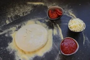 Pizzateig auf bemehlter Arbeitsfläche mit Schüsseln Peperoni, geriebener Mozzarella und Tomatensoße