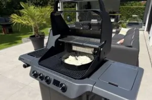 Gasgrill mit zwei Grillebenen, auf der unteren Ebene eine Gusseisenpfanne mit gebratenen Zwiebeln für Whiskyzwiebeln, auf der oberen Ebene Grillrost, im Garten aufgestellt