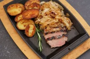 Gegrilltes Rumpsteak medium-rare geschnitten, mit karamellisierten Whiskyzwiebeln und knusprigen Röstkartoffeln auf schwarzem Tablett, garniert mit Rosmarin