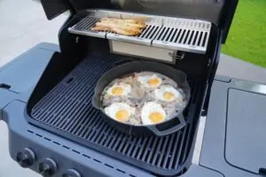 Gusseisenpfanne mit fünf Spiegeleiern auf gegrilltem Spargel auf dem Grill, im Hintergrund gegrilltes Hähnchen auf der oberen Grillplatte