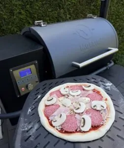 Pizza mit Salami und Pilzen auf schwarzem Grillrost eines Pelletgrills mit digitaler Temperaturanzeige im Hintergrund