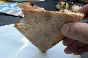 Angeschnittenes Stück Steinofenpizza mit krosser, goldbraun gebackener Kruste und dünn belegter Oberfläche, in Hand gehalten